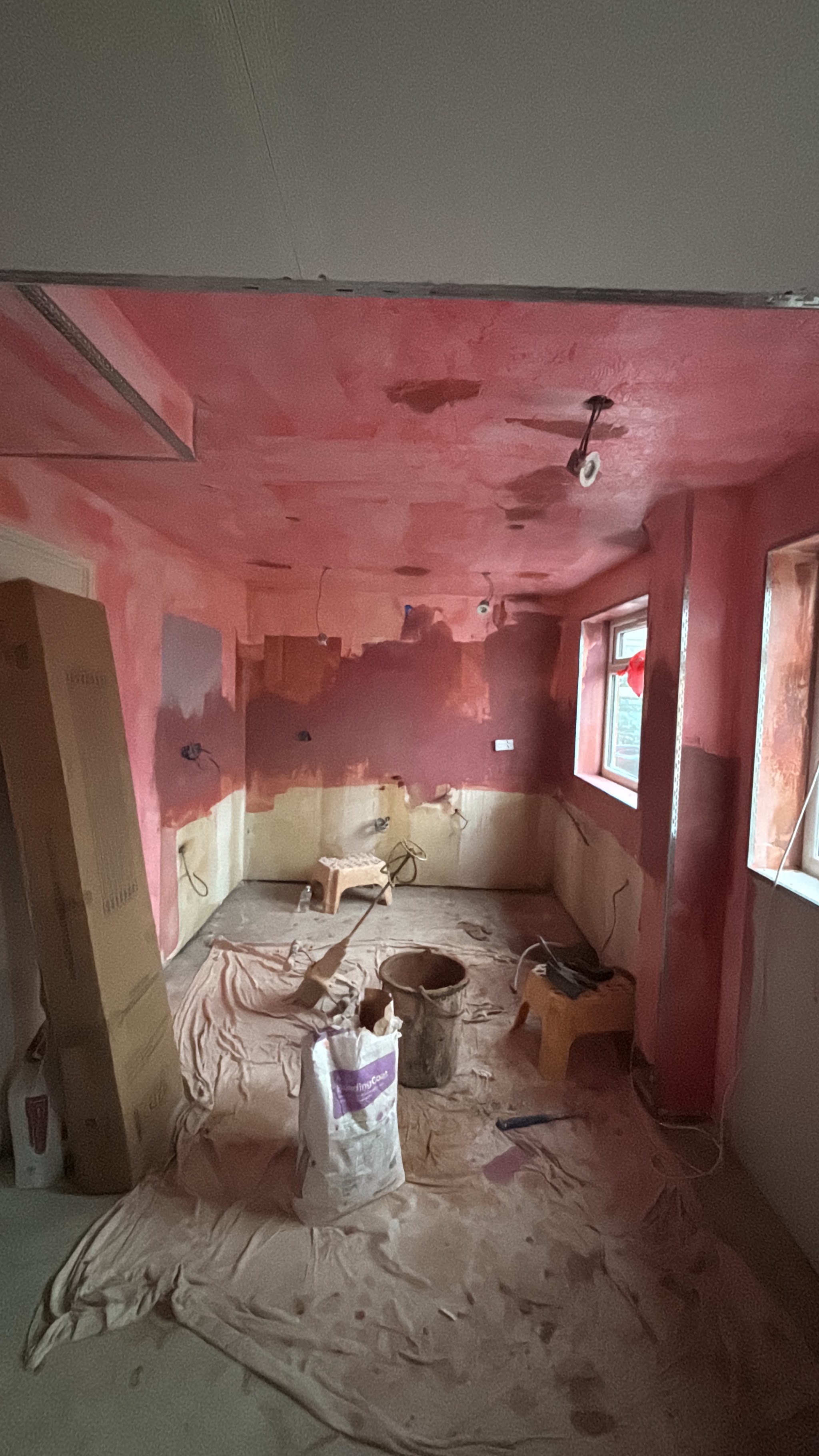 A kitchen mid-job with the walls and ceiling primed pink, dust sheets down and a plaster bag ready